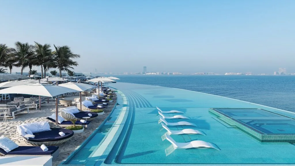 Piscine à débordement luxueuse avec vue sur l’océan, entourée de transats et palmiers.